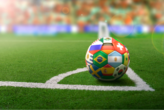 Bola de futebol posicionada em um campo, próxima à linha do escanteio. O destaque está no design da bola, composta por bandeiras de diversos países. Ao fundo, vê-se parte do estádio desfocado, com arquibancadas coloridas e iluminação suave.