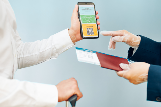 Pessoa mostrando um celular com um QR code para um agente que segura um passaporte e um cartão de embarque, durante um procedimento de checagem de viagem