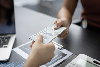Pessoa entregando notas de dólar a outra em um ambiente de escritório. Sobre a mesa, há documentos com gráficos, um laptop e uma calculadora