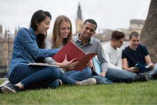 Grupo de jovens sentados em um gramado, estudando juntos ao ar livre. Eles folheiam livros e sorriem. Ao fundo, há construções históricas que remetem a uma cidade europeia.