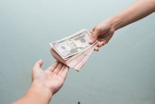 Duas mãos aparecem em destaque, uma entregando e outra recebendo um maço de notas de dólar americano